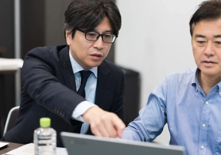 スタッフがノートPCの画面を指さしながら隣の人物に説明している様子