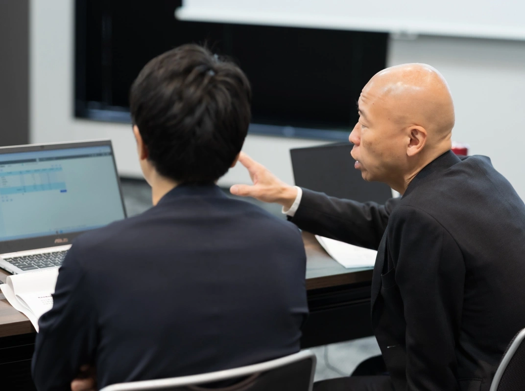 担当者がノートPCの画面を指し示しながら隣の人物に説明している様子