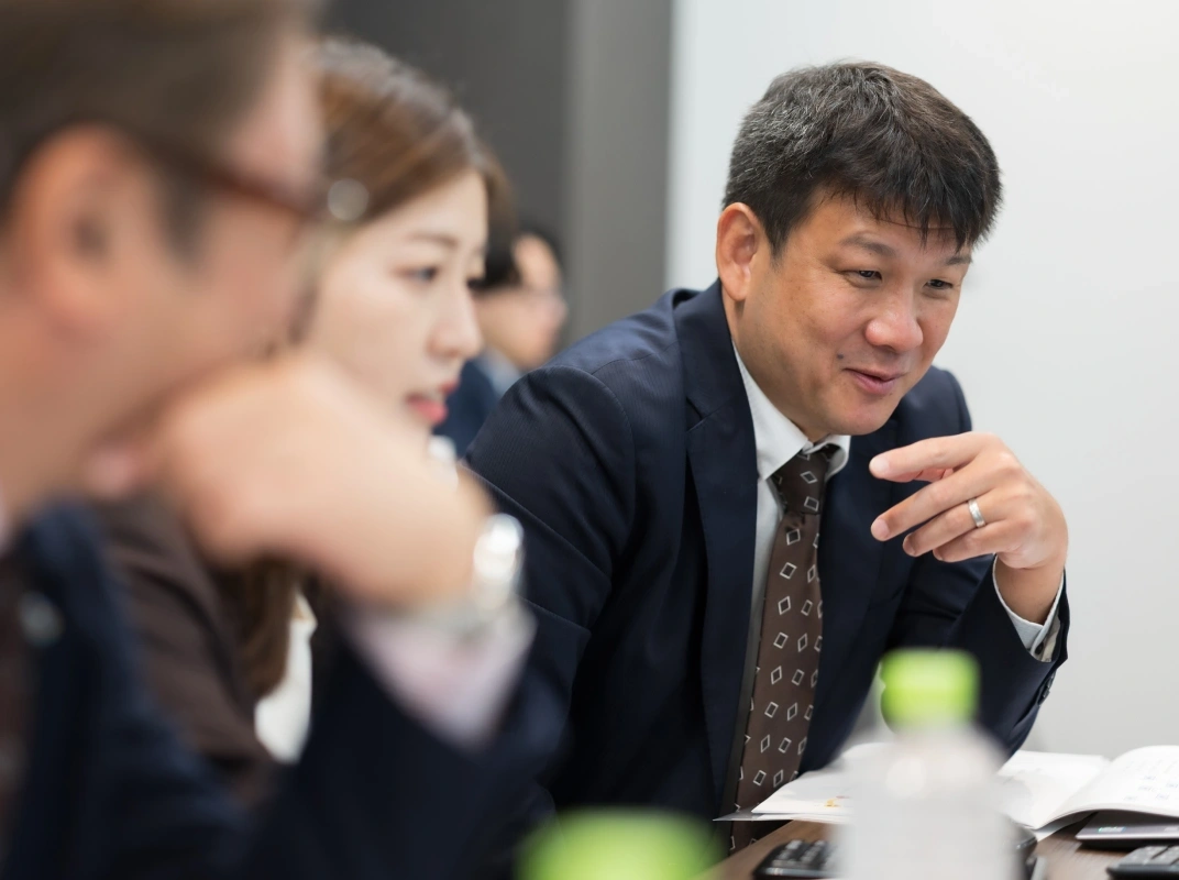 会議室で複数名が向かい合い、スーツ姿の男性が手振りを交えながら話している様子